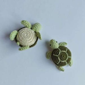 May include: Two handmade crochet sea turtles. One turtle has a cream-colored shell with a dark green border and light green flippers. The other turtle has a dark green shell with light green flippers. Both turtles have black eyes.
