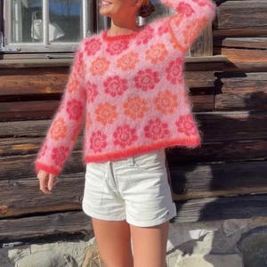 Puede incluir: Un suéter rosa y difuso con un estampado floral en tonos de rosa y naranja. El suéter tiene cuello redondo y mangas largas. La modelo lleva shorts blancos.