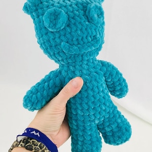 May include: A turquoise plush doll with a smiling face, round eyes, and small arms and legs. The handmade doll is held in a person's hand. The doll is made of a soft, textured material.