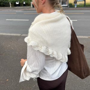 May include: Cream-colored knitted shawl with a ruffled edge. The shawl is worn over a white button-down shirt with thin vertical stripes. A brown tote bag is visible.