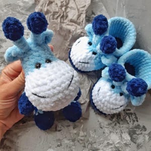 Può includere: Giraffa e scarpine per neonati all'uncinetto nei toni del blu e del bianco. La giraffa ha dettagli blu scuro sulle orecchie, sui piedi e sulle corna. Le scarpine hanno un design simile, con un volto sorridente e dettagli blu scuro.