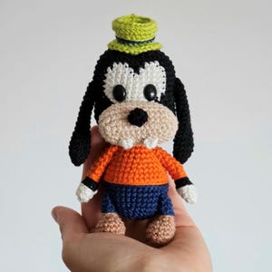 May include: A handmade crochet figure of a cartoon dog character. The figure has a black and white face, black ears, an orange shirt, blue shorts, and a green hat. The figure is being held in a person's hand.