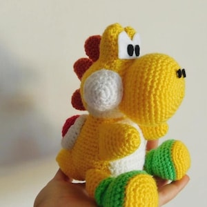 Può includere: Bambola Yoshi all'uncinetto, gialla, bianca, rossa e verde. Il peluche presenta un corpo arrotondato, grandi occhi e una sella. La bambola è un personaggio popolare della serie di videogiochi Super Mario.