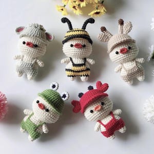 Pode incluir: Cinco bonecas de crochê feitas à mão, cada uma vestida como um animal diferente. As bonecas são brancas com bicos vermelhos e olhos pretos pequenos. Elas usam chapéus e macacões em várias cores, incluindo cinza, amarelo, verde, vermelho e bege.