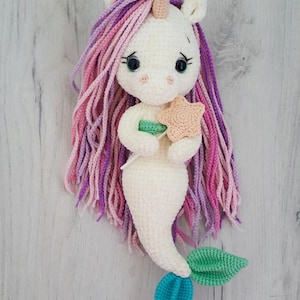 May include: A handmade crochet mermaid unicorn doll. The doll has a white body, a cream-colored horn, and long, flowing hair in shades of pink and purple. It holds a tan starfish and a green wand. The mermaid tail is teal and green.