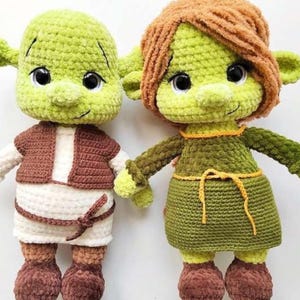 May include: Two handmade crochet dolls, one resembling a green ogre with a brown vest and white shirt, and the other a green ogre with a green dress and orange hair. Both dolls have large black eyes and are made with soft yarn.