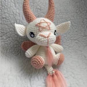 May include: A crocheted goat doll with white and peach-colored yarn. The doll features large, curved horns, a five-pointed star on its forehead, and a fluffy tail. The goat has black eyes and small wings.