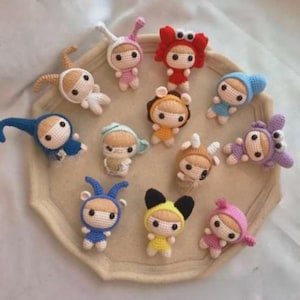 May include: A collection of handmade crochet dolls, each dressed in unique animal or character costumes. The dolls feature various colors, including blue, yellow, pink, and brown, with detailed facial features and costume elements. They are arranged on a light-colored tray.