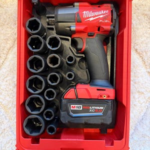 May include: A red Milwaukee FUEL impact wrench kit in a red plastic case. The kit includes the impact wrench, a battery, and a selection of black sockets. The case has a clear lid and is designed for tool storage and transport.