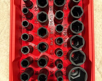 May include: A red plastic storage tray containing a collection of black metal impact sockets of various sizes. The tray features molded slots with printed labels for each socket, including fractional inch and metric measurements.