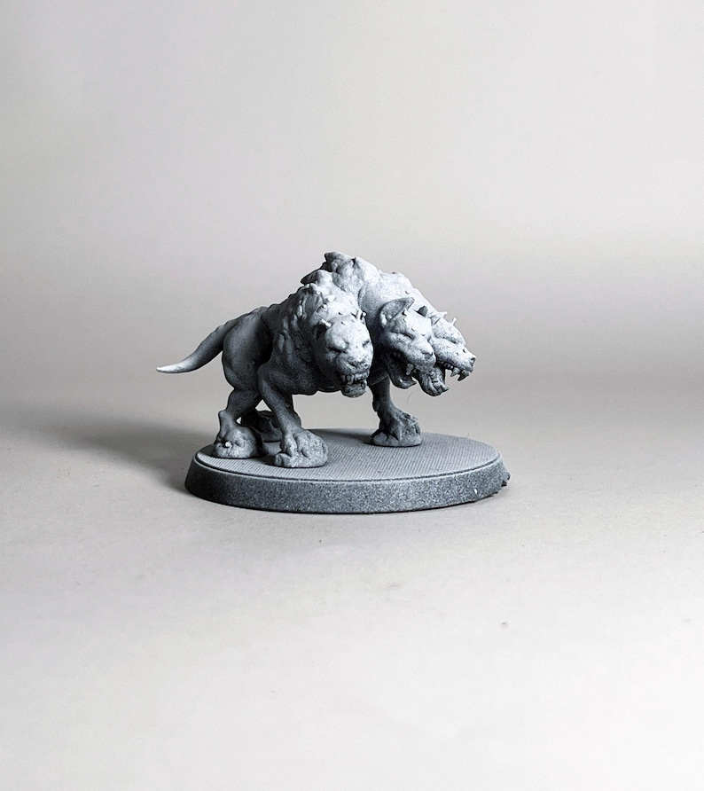 May include: A gray, three-headed Cerberus figurine, standing on a circular base. The mythical creature has a lion-like body with three snarling heads and is sculpted in a detailed, realistic style. The figurine is a fantasy-themed collectible.