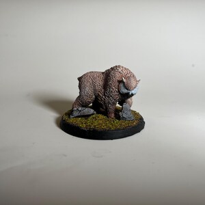 May include: A miniature figurine of a brown, furry beast with curved horns and a white face. The creature is posed on a circular base with green moss and small rocks. The base has a black border.
