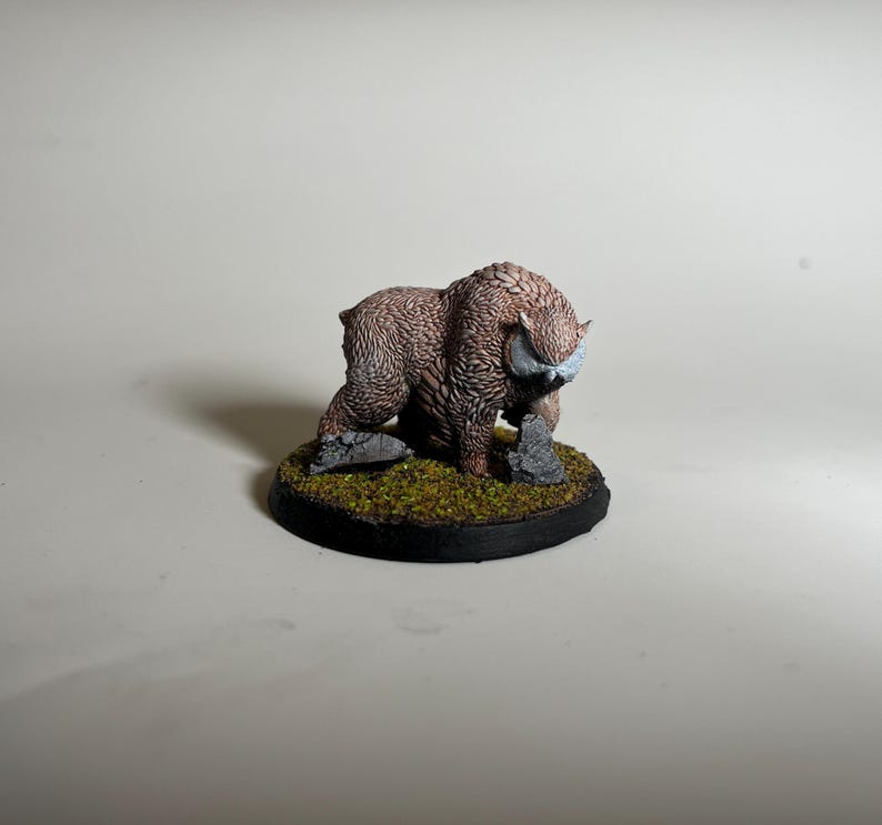 May include: A miniature figurine of a brown, furry beast with curved horns and a white face. The creature is posed on a circular base with green moss and small rocks. The base has a black border.