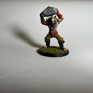 May include: A miniature figurine of a muscular warrior, holding a gray rock overhead. The figure is painted with detailed features, including a beard, red accents, and green and yellow clothing. The base is a circular platform with green textured material.