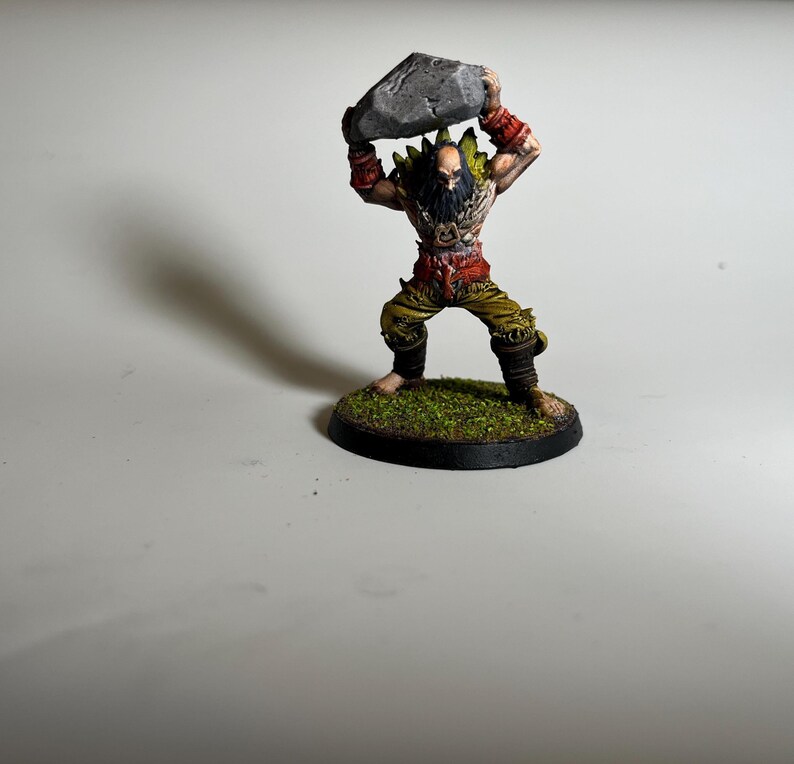 May include: A miniature figurine of a muscular warrior, holding a gray rock overhead. The figure is painted with detailed features, including a beard, red accents, and green and yellow clothing. The base is a circular platform with green textured material.