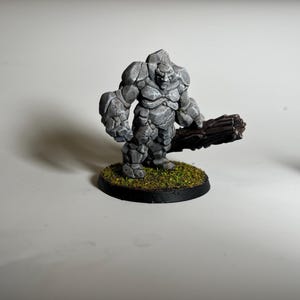 May include: A gray stone golem miniature figure with a textured surface, holding a brown log. The figure stands on a circular base covered in green and brown faux grass. The base has a black border.