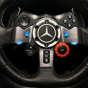 May include: A black leather-wrapped racing steering wheel featuring a central Mercedes-Benz logo, blue L2, R2, L3, and R3 buttons, a red dial, a directional pad, PlayStation action buttons, and a center console with Share, Option, and PS buttons.