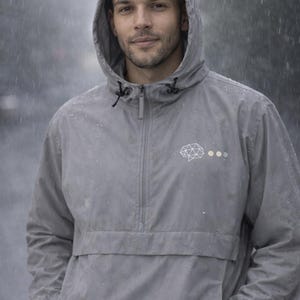 Könnte beinhalten: Graue Kapuzen-Anorakjacke mit halbem Reißverschluss und Vordertasche. Die Jacke hat ein weißes geometrisches Gehirnlogo und drei kleine Punkte. Die Person trägt die Jacke im Regen.