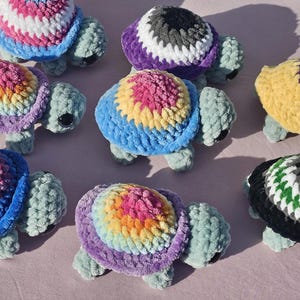 May include: A selection of handmade crochet turtles in various colours. Each turtle features a textured shell in shades of blue, pink, yellow, purple, and green. The turtles have light green bodies and black eyes, creating a whimsical and colourful display.