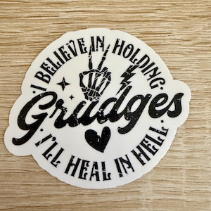 May include: A white sticker with black text and graphics. The text reads "I BELIEVE IN HOLDING Grudges I'LL HEAL IN HELL." A skeleton hand forms a peace sign. A heart and lightning bolt are also featured.