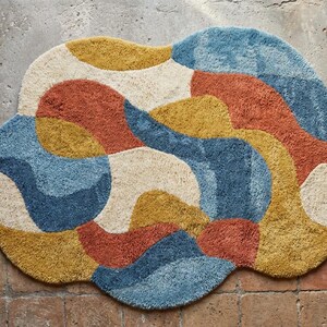 May include: A colorful, abstract rug with a cloud-like shape. The rug features a mix of colors, including blue, orange, yellow, and cream. The rug is made of a soft, textured material and is placed on a brick and concrete surface.