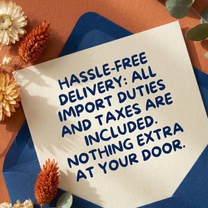 May include: A cream-colored card with blue text that reads "HASSLE-FREE DELIVERY: ALL IMPORT DUTIES AND TAXES ARE INCLUDED. NOTHING EXTRA AT YOUR DOOR." The card is inside a blue envelope, with dried flowers and greenery.