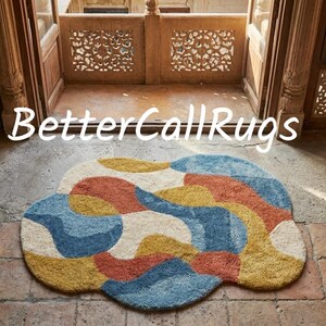 May include: A colorful, abstract rug with a cloud-like shape. The rug features a pattern of blue, yellow, orange, and cream shapes. The text "BetterCallRugs" is displayed above the rug.
