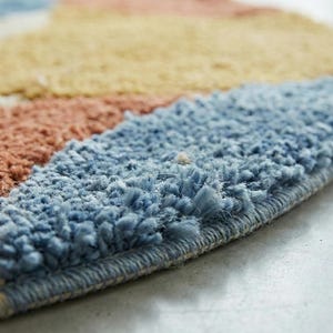 May include: Close-up of a colorful, round rug with a textured surface. The rug features a design with stripes of blue, peach, yellow, and white. The rug's edge is visible, showing a tightly woven border.
