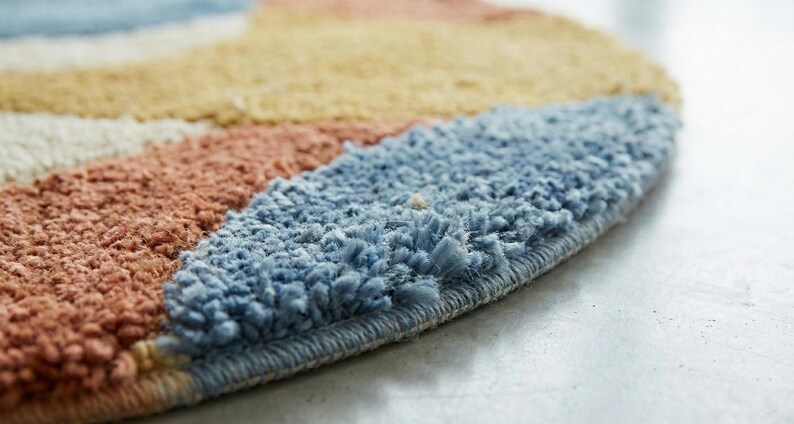 May include: Close-up of a colorful, round rug with a textured surface. The rug features a design with stripes of blue, peach, yellow, and white. The rug's edge is visible, showing a tightly woven border.