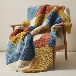 May include: A colorful, patterned throw blanket draped over a wooden chair. The blanket features large, irregular shapes in shades of blue, orange, yellow, and cream. The chair has a light wood frame and a white seat.