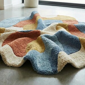 May include: A colorful, abstract rug with a wavy, irregular shape. The rug features patches of blue, peach, yellow, and cream colors. The rug is made of a soft, textured material and is placed on a gray floor.