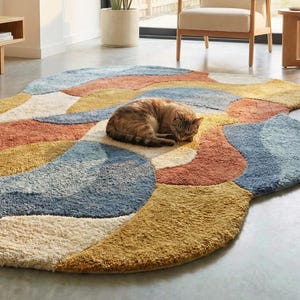 May include: A colorful, abstract area rug with a soft, textured surface. The rug features a pattern of overlapping shapes in shades of blue, yellow, orange, and cream. A cat is curled up asleep on the rug. Home decor.