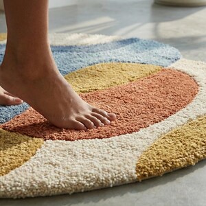 May include: A colorful, round bath mat with a textured surface. The mat features a design with sections of blue, orange, yellow, and cream colors. The image shows a person's feet standing on the mat.