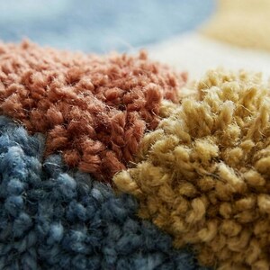 May include: Close-up of a colorful rug with a textured, looped pile. The rug features sections of blue, brown, and yellow, creating a geometric pattern. The soft, plush texture is visible in the detailed shot.