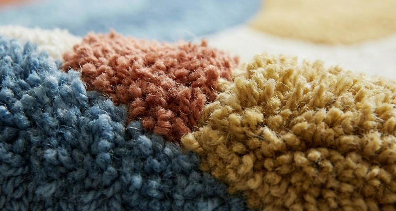 May include: Close-up of a colorful rug with a textured, looped pile. The rug features sections of blue, brown, and yellow, creating a geometric pattern. The soft, plush texture is visible in the detailed shot.