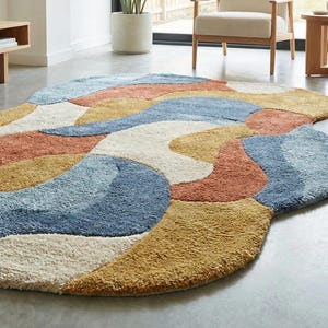 May include: A large, abstract area rug with a soft, textured surface. The rug features a flowing design with sections of cream, rust, blue, and gold. The rug is placed on a gray concrete floor, adding a modern touch to the room.