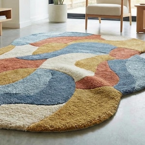 May include: A large, abstract area rug with a soft, textured surface. The rug features a flowing design with sections of white, blue, orange, and gold. The rug is placed on a gray floor in a modern living space.