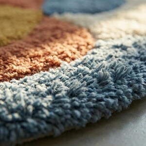 May include: Close-up of a colorful, textured rug. The rug features a pattern of overlapping shapes in shades of blue, brown, orange, and yellow. The rug's surface has a soft, plush texture, suggesting a comfortable and inviting feel.