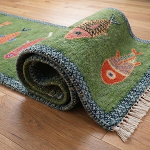 May include: A green runner rug featuring a repeating pattern of stylised fish in shades of orange, pink, and purple. The rug has a dark blue textured border and white fringe at the ends. The material appears to have a thick, tufted pile texture.