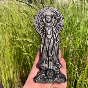 Może przedstawiać: Ciemnoszara statua kobiety z okrągłą aureolą, stojąca na szczegółowej podstawie. Statua trzymana jest w dłoni na tle zielonej trawy. Kobieta przedstawiona jest w powłóczystej szacie.