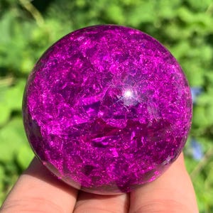 May include: A spherical object made of clear glass with a vibrant magenta crackle texture inside. The surface is smooth and reflective, showing internal fractures that create a multifaceted appearance throughout the entire round shape.