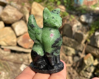 May include: A carved stone figurine of a winged cat, crafted from green zoisite with black hornblende inclusions and a small pink ruby spot on the chest. The cat is depicted in a seated position with wings extended upward, featuring a smooth, polished finish.