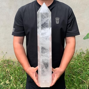 Könnte beinhalten: Ein großer, klarer, sechsseitiger Quarzkristall mit einer spitzen Oberseite und einer flachen Basis. Der Kristall ist transparent mit sichtbaren inneren Rissen und wolkigen Einschlüssen in seiner länglichen, hexagonalen Säulenstruktur.