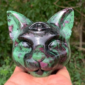 May include: A carved stone cat head figurine featuring a polished surface with a mottled pattern of deep green and vibrant magenta. The sculpture includes a third eye detail on the forehead, pointed ears, and defined facial features.