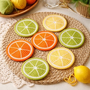 May include: A collection of handmade crochet coasters in the shape of citrus fruit slices. The coasters feature bright colors including orange, yellow, and green, with white detailing. The coasters are arranged on a woven placemat, creating a vibrant and summery aesthetic.