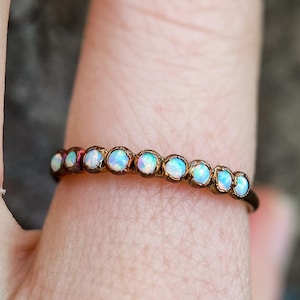 May include: A delicate ring featuring a row of small, round opals set in a thin, bronze-colored band. The opals display a range of iridescent colors, including white, blue, and hints of pink. The ring is shown on a finger.
