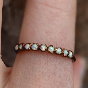 May include: A delicate ring featuring a row of small, round opals set in a thin, dark metal band. The opals have a milky white color and are evenly spaced around the ring. The ring is shown on a finger.