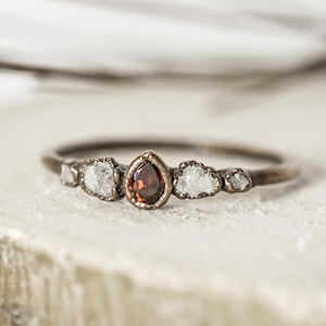 May include: A delicate ring with a central teardrop-shaped, reddish-brown gemstone, flanked by three smaller, irregular, white gemstones. The ring is set in a thin, bronze-colored band. The ring is displayed on a light-colored, textured surface.