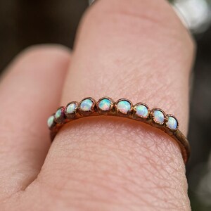 May include: A delicate gold ring adorned with small, iridescent opals. The ring features a thin band and a series of opals set closely together, creating a subtle yet eye-catching design. The opals display a range of colors, adding a touch of elegance.