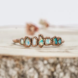 Peut inclure: Une bague de couleur cuivre ornée d'une rangée de pierres d'opale bleu clair et blanches de forme irrégulière. La bague est posée sur une surface en bois, avec un arrière-plan flou. Les pierres d'opale ont un aspect naturel unique.